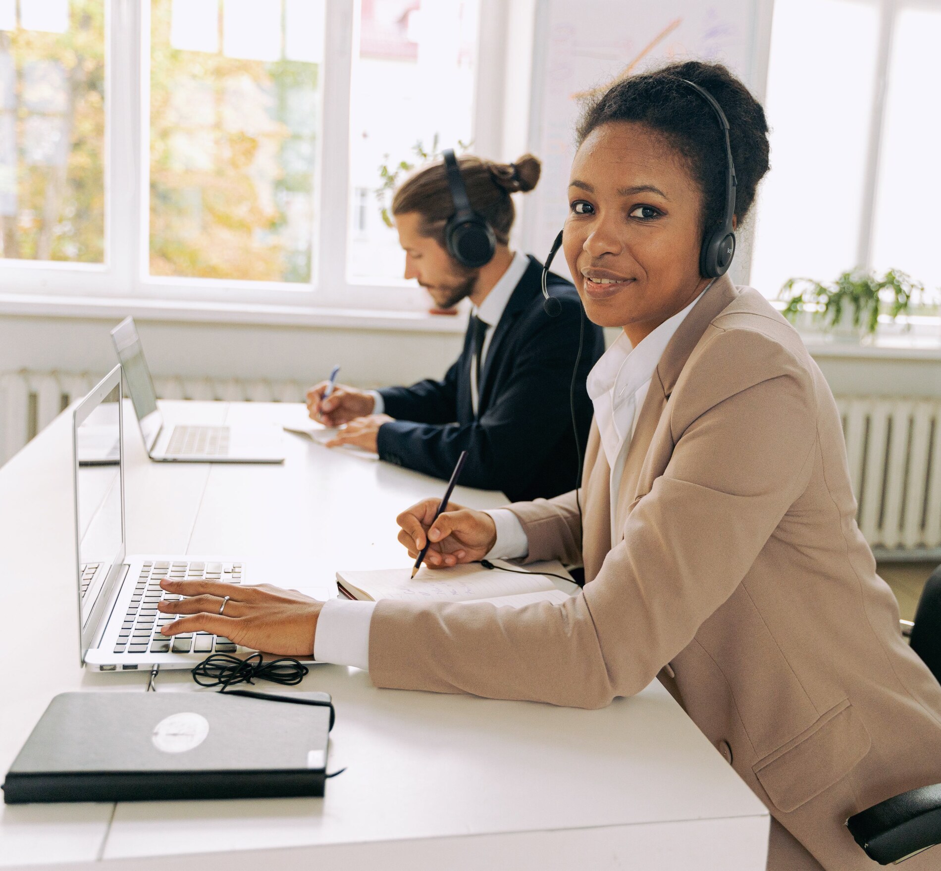 A woman wearing a headset taking a call at an office desk, with a colleague on a call behind her