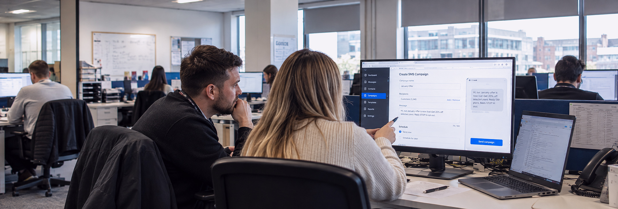 Two colleagues in an open-plan office setting up an SMS campaign on a desktop screen