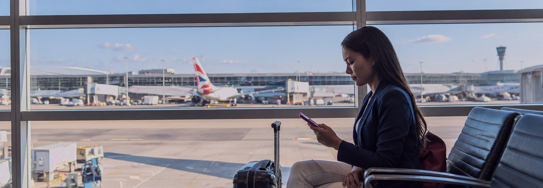 A woman in an airport departure lounge checking her phone, with aircraft on the apron beyond the window